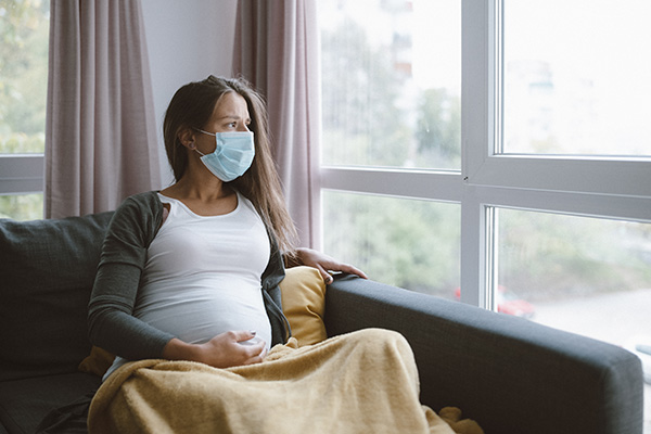 worried pregnant woman sitting at home with protective face mask and looking through the window.